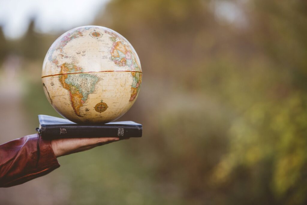A person’s hand holding up a Bible atop which sits a globe.